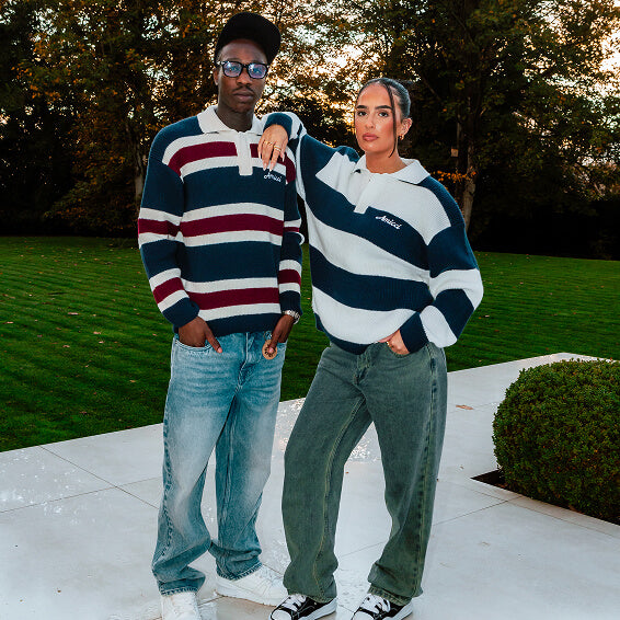 A man and a woman standing outdoors on a patio in front of a grassy lawn and trees, both wearing matching striped rugby-style sweaters with navy, white, and burgundy horizontal bands. The man wears blue jeans and white sneakers, and the woman wears olive green jeans and black-and-white sneakers.